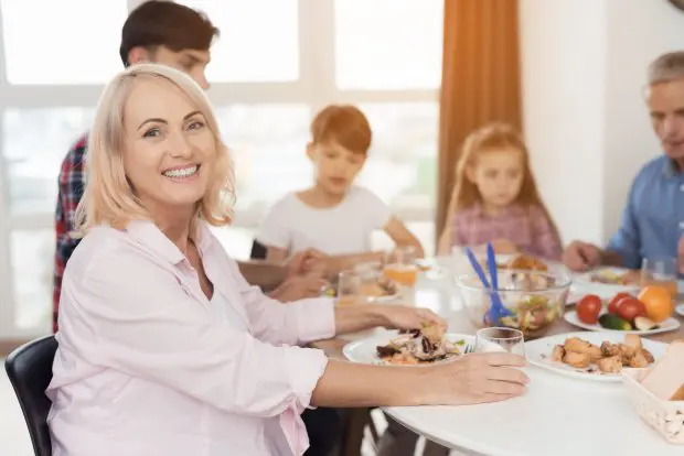 A middle-aged woman posing against the backdrop of her family, who sits at the Thanksgiving table