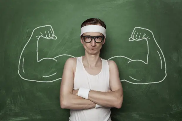 Young adult male that is in front of a chalkboard and that is skinny. On the chalkboard behind him is a drawing of very strong arms stemming from his body.