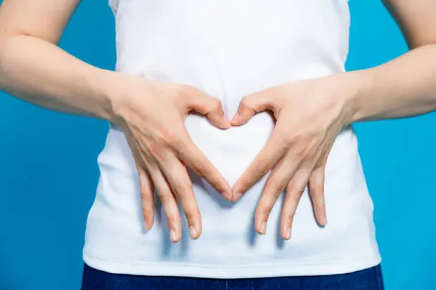 View of a woman's stomach and she is making a heart with her hands and holding them over her belly on top of her white shirt.