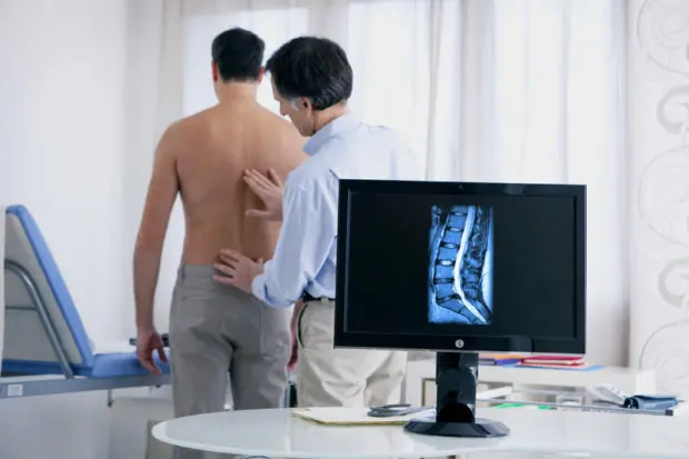 A shirtless patient in a medical office that is having his back examined. On the screen behind him, there is an x-ray image of a bulging disc in his back.