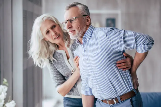 An elderly couple. The woman is helping the man to stand, as he is experiencing back pain that has him bending forward.