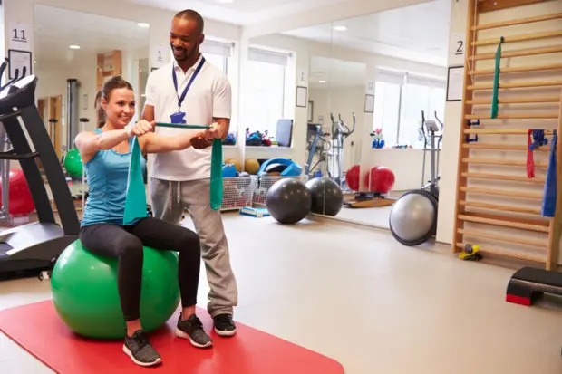 A young adult female that is doing arm exercises for rehabilitation. She is sitting on an exercise ball and is getting help from a chiropractic physical therapist.