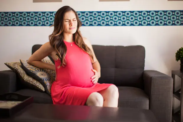 A very pregnant brunette woman that is experiencing lower back pain. Her face is one of pain.