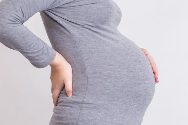 A close-up view of a very pregnant woman's torso. She has one hand on her lower back and another hand on her belly.