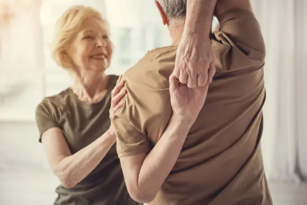 An elderly woman helping her companion as he does a stretch behind his back that is hard to do. He is hooking both hands together behind his back, with one arm over his head and the other arm down at his side.