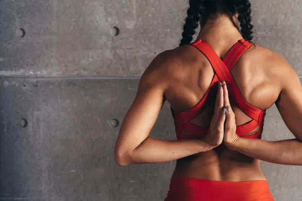 An athletic woman that is doing a stretch behind her back, putting her arms together as she faces a wall.
