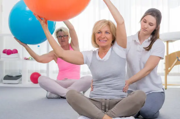 A middle-aged woman that is doing stretches with a ball in-office with a medical professional.