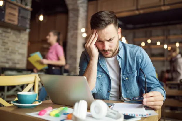A man that is studying in a coffee-shop type of establishment. He is taking a minute away from his computer and studies to hold his head that is in pain.