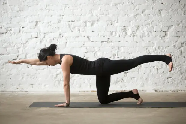 A woman that is doing yoga in her home. She is dressed in black as she is doing yoga in front of a white wall.