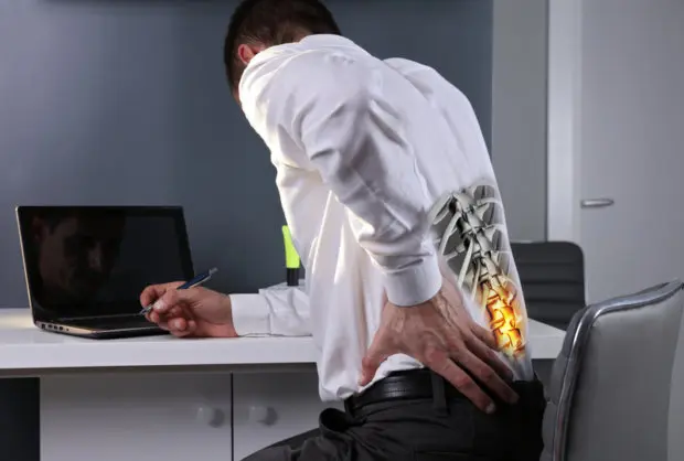 A young adult male that is holding his lower back in pain as he sits at an office desk working. A digital image of his lower back and spine is showing through his shirt. 