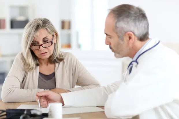 An middle-aged woman with shoulder-length hair that is speaking with a medical professional as he is references things on his clipboard in front of him.