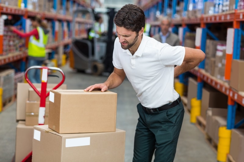 A man that is standing in a warehouse with various boxes in front of him. He is holding his back to suggest he hurt his back from lifting.
