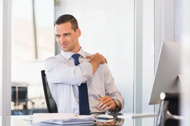 An attractive, young male that is in business attire and holding his shoulder as he is experiencing upper back pain during work.