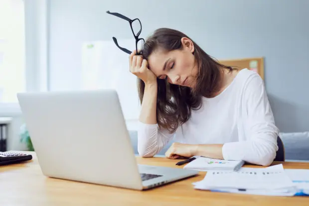 A woman that is tired while she is working on a laptop computer. She has pulled off her glasses and is laying her head on her hand.
