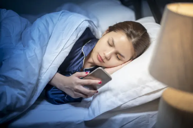 A young adult woman that is on her cell phone while she is in bed for nighttime.